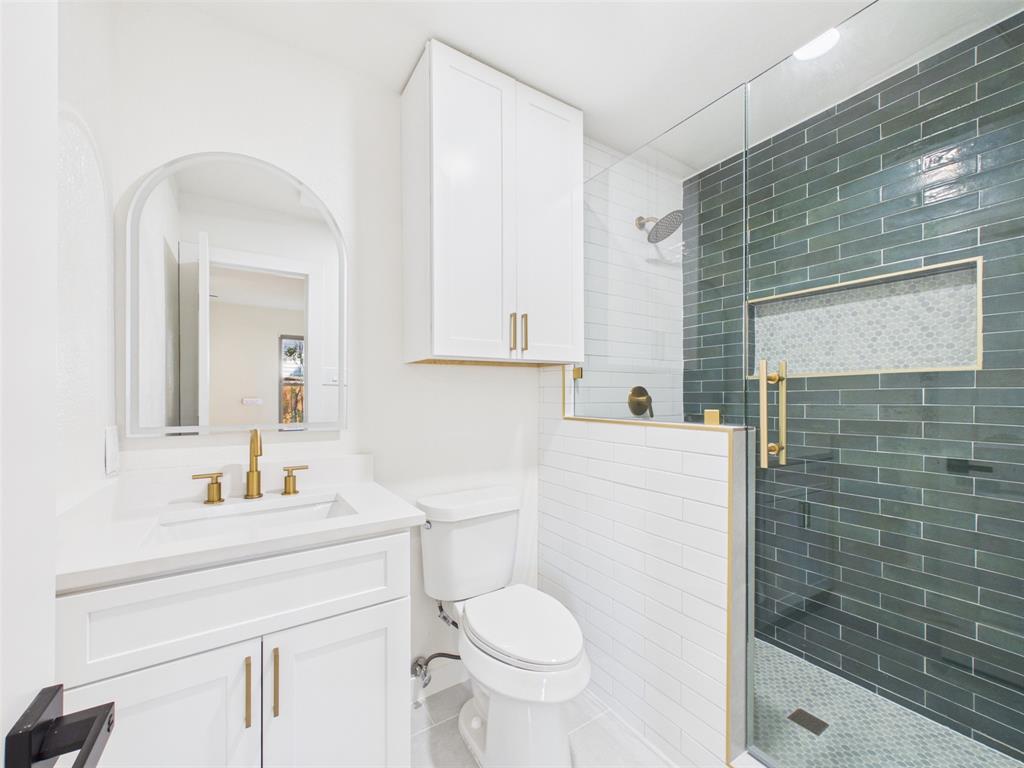 Bathroom featuring a white vanity with gold-tone hardware, arched wall mirror, walk-in shower with dark horizontal tile, built-in shower niche, and glass enclosure