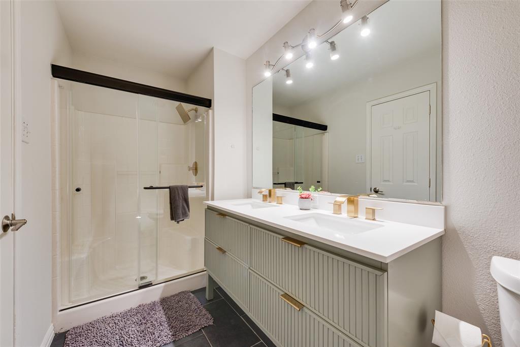 Double vanity featuring a fluted-style cabinet base, stone countertop, integrated rectangular sinks, and metallic finish hardware, complemented by a walk-in shower with a glass enclosure