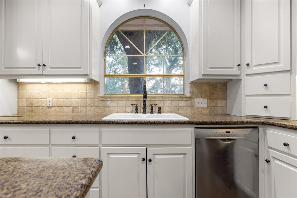 Kitchen featuring white shaker-style cabinetry, stone-finish countertops, textured tile backsplash, arched window with decorative muntins, and a dual-basin white sink with a dark-finish faucet