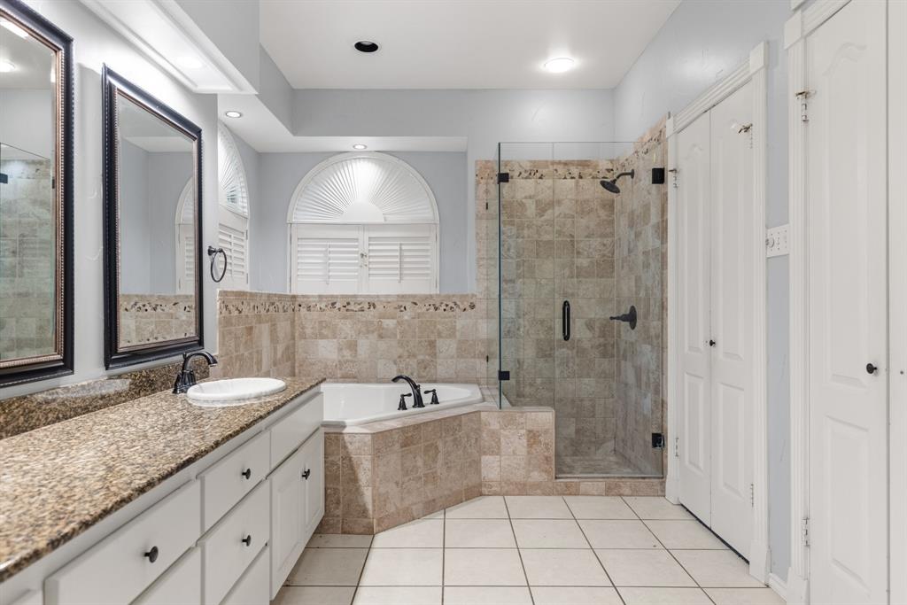 Dual-sink vanity with stone countertop, corner soaking tub with tile surround, glass-enclosed walk-in shower, and arched window with plantation shutters