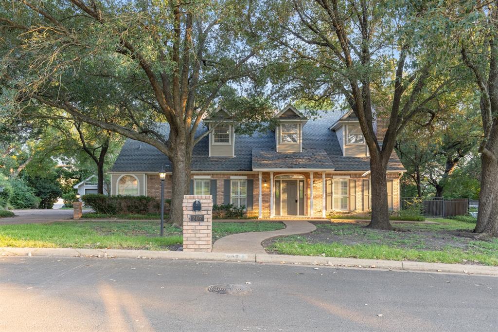Two-story brick exterior featuring a steeply pitched roof, dormer windows, covered front entrance with columns, and mature canopy trees