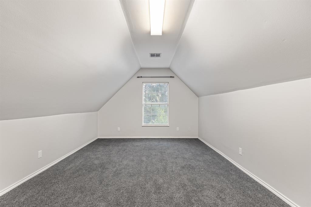 Vaulted ceiling featuring neutral wall paint, gray carpeting, a centrally located linear flush-mount light fixture, and a single window with blinds