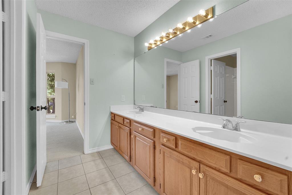 Dual-sink vanity with wood-finish cabinetry, solid-surface countertop, full-width mirror, and multi-bulb light fixture, complemented by tiled flooring