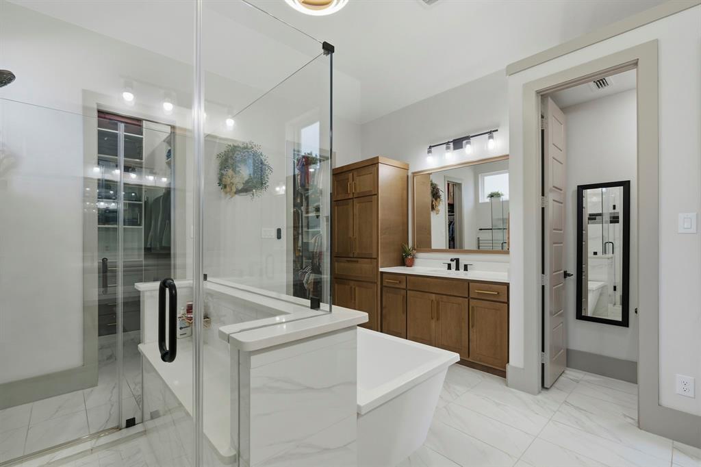 Bathroom featuring large-format tile flooring, a glass-enclosed shower, a soaking tub with stone-finish surround, built-in wooden cabinetry, and a vanity with a rectangular sink and matching hardware