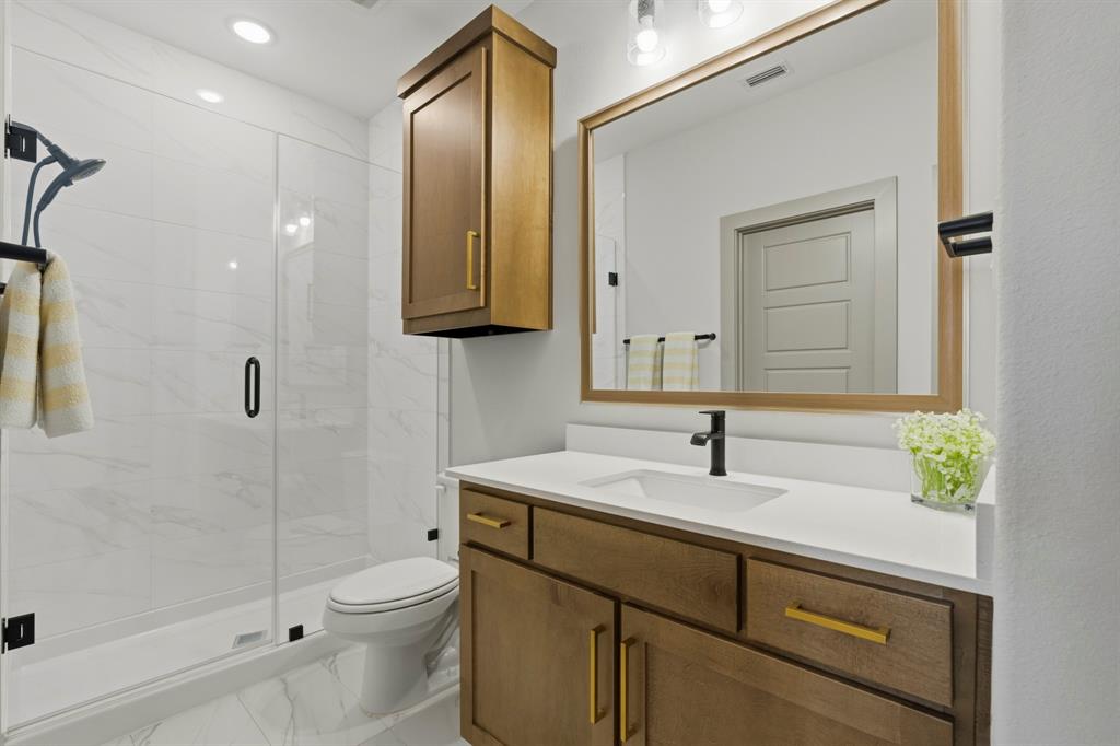 Bathroom featuring wood-finish cabinetry with brass hardware, quartz countertop with integrated sink, walk-in shower with glass enclosure, and large-format tile flooring
