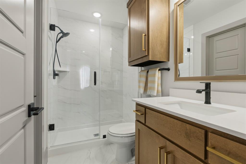 Bathroom featuring a glass-enclosed shower with large-format wall tile, wood-finish vanity cabinetry with gold-toned hardware, quartz-composite countertop, and matte black plumbing fixtures