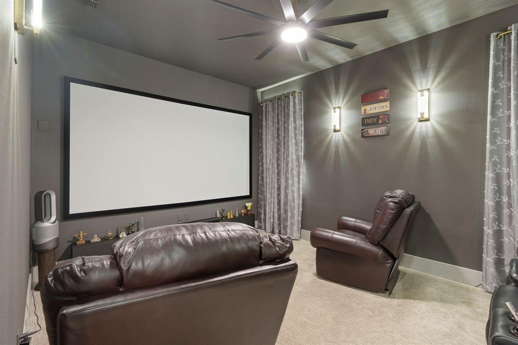 Media room featuring a fixed projection screen, neutral carpeting, a multi-blade ceiling fan with integrated lighting, and dual wall-mounted light fixtures