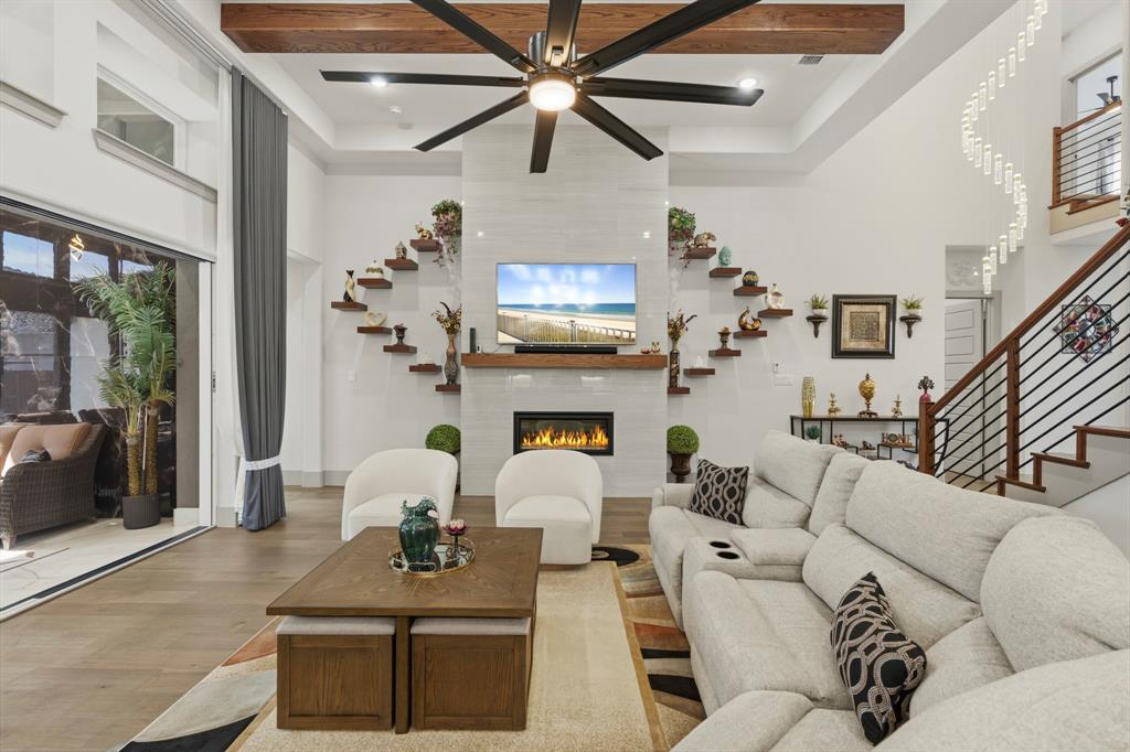High-ceilinged living area featuring a fireplace with a full-height tile surround, exposed wooden beam, large-scale multi-blade ceiling fan, and staircase with metal railing