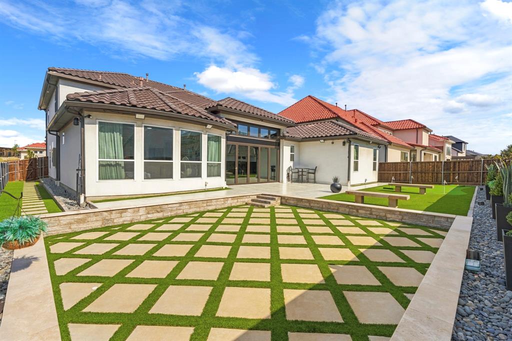 Rear exterior featuring a multi-level terrace with geometric stone pavers and synthetic turf inlay, retaining walls, covered patio, and tile roof