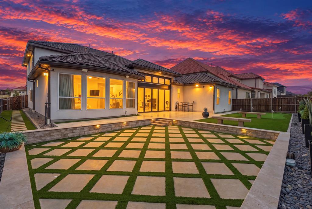 Rear exterior featuring a tile roof, geometric paver patio with turf inlays, raised stone retaining walls with integrated landscape lighting, and a synthetic turf lawn