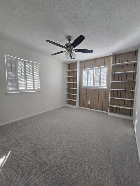 Room featuring wall-to-wall carpeting, ceiling fan, wood-paneled accent wall with built-in shelving, and white plantation-style shutters