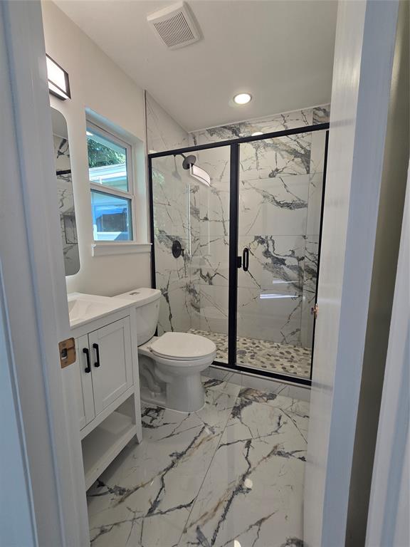 Bathroom featuring large-format patterned tile flooring, a walk-in shower with black-framed glass enclosure, stone-finish wall tile, pebble-style shower floor, and a white cabinet vanity