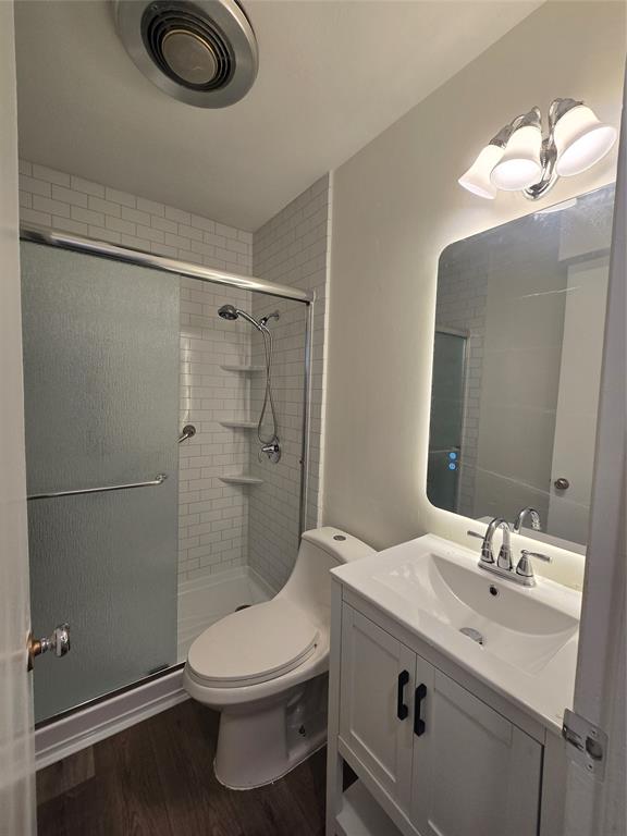 Bathroom featuring a white vanity with an integrated sink, a rectangular mirror with integrated perimeter lighting, a walk-in shower with white subway tile surround, and wood-finish flooring