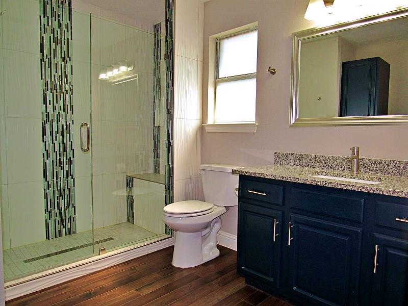 Walk-in shower with vertical mosaic tile accents and glass door, navy blue vanity cabinet, stone-finish countertop, and wood-finish flooring