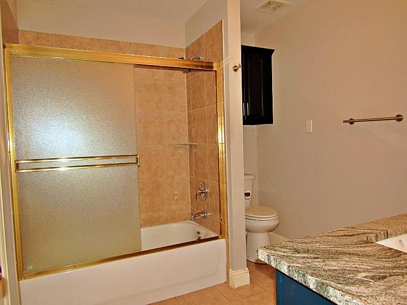 Bathroom featuring a tub-shower combination with floor-to-ceiling tile surround, gold-toned sliding glass enclosure, stone-surface countertop, and built-in dark wood-finish cabinetry