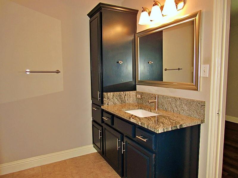 Dark-toned cabinetry featuring a tall built-in linen storage tower, stone-surface countertop with an integrated rectangular sink, three-light fixture with frosted glass shades, and a framed mirror