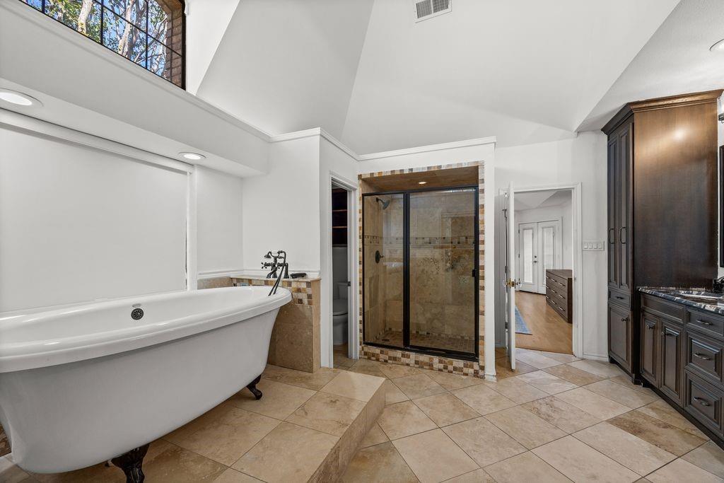 Bathroom featuring a high vaulted ceiling, freestanding clawfoot tub on a raised tile platform, enclosed glass-door shower with mosaic tile accents, dark-finish wood cabinetry, and large-format tile flooring