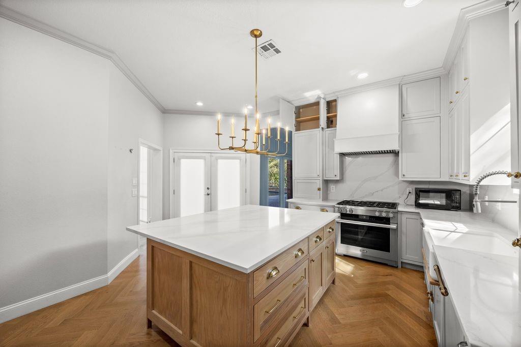 Kitchen featuring a herringbone wood-finish floor, a center island with a stone countertop, white cabinetry with matching range hood, stainless steel gas range, and a brass-finish chandelier