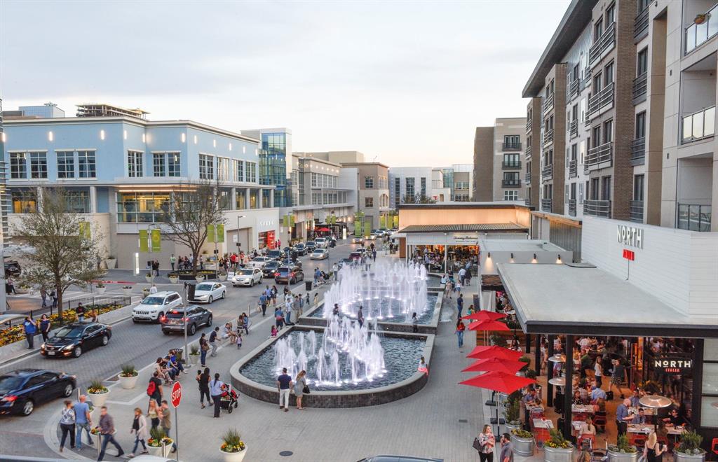 Central urban development featuring multi-story buildings with residential and commercial spaces, a prominent water fountain feature, pedestrian walkways, and street-level dining establishments