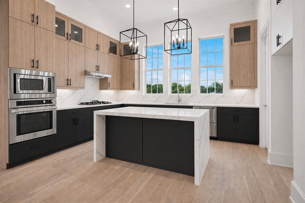Contemporary kitchen featuring a waterfall island with a stone countertop, light wood-finish flooring, stainless steel appliances, and dual black geometric pendant lights