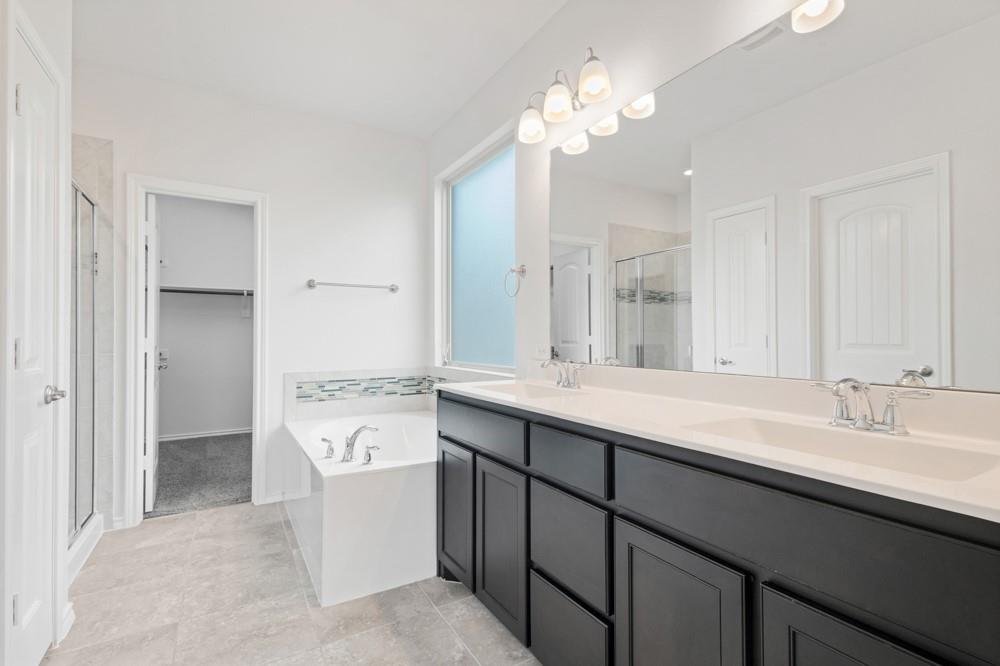Dual-sink vanity with dark cabinetry, light countertops, soaking tub with tile backsplash, and full-width mirror