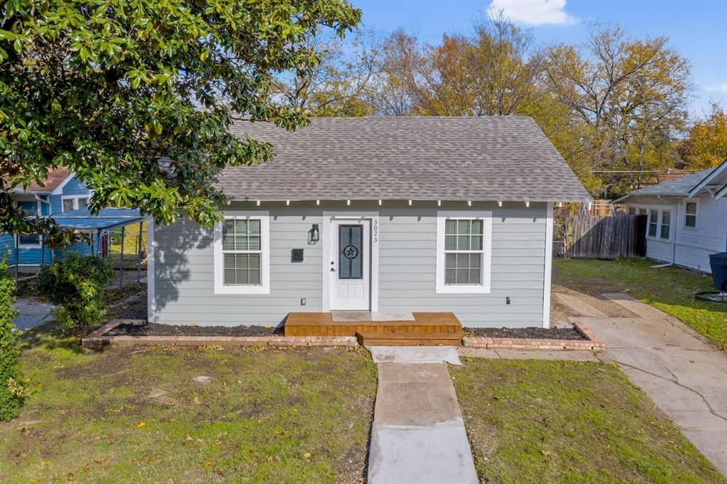 Single-story exterior featuring light gray horizontal siding, asphalt shingle roofing, a wood-plank front porch, and brick-edged garden beds