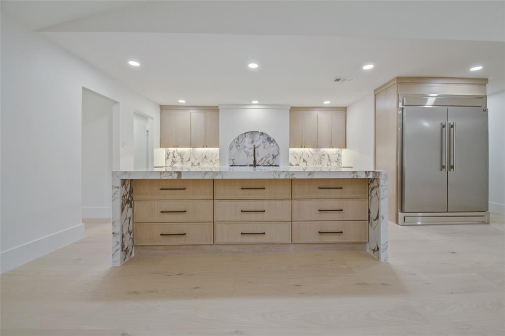 Large kitchen island featuring light-finish cabinetry, a waterfall-edge countertop, stone backsplash with arched niche, recessed ceiling lighting, and integrated stainless steel refrigerator