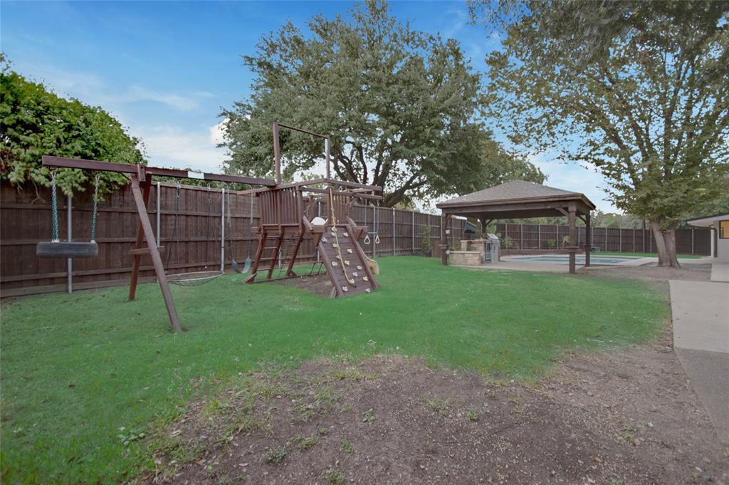 Backyard featuring a grass lawn, wooden perimeter fencing, built-in wooden play structure, and a covered pavilion with stone-base outdoor kitchen