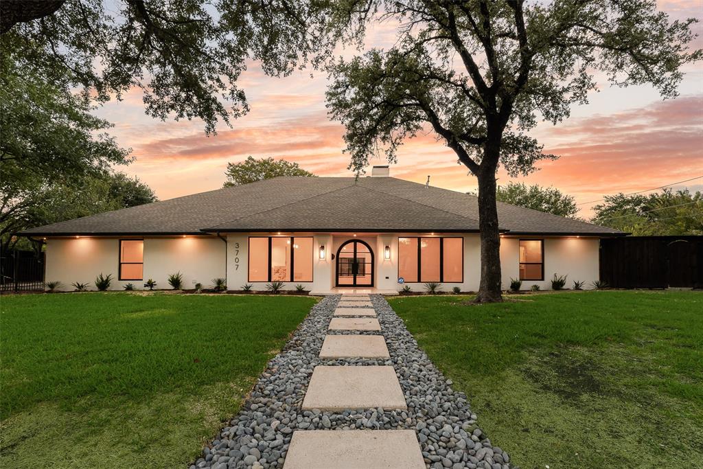 Single-story stucco exterior featuring an arched double-door entryway, large-pane windows, a stone-accented concrete paver walkway, and mature landscape trees