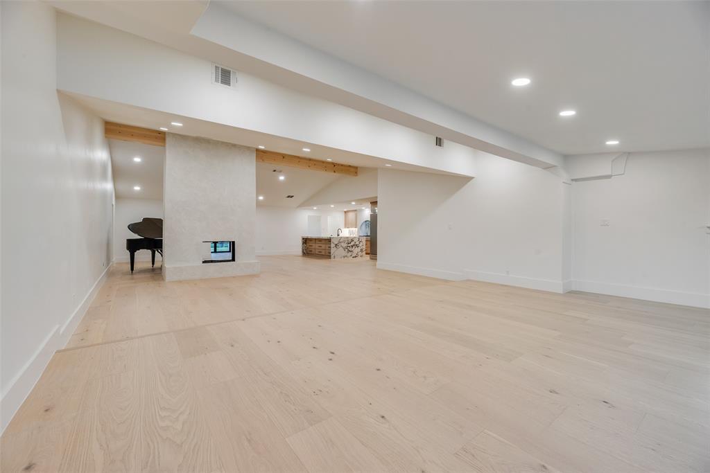 Open-concept interior featuring light wood-finish flooring, recessed lighting, exposed wood ceiling beams, and a centrally positioned masonry fireplace