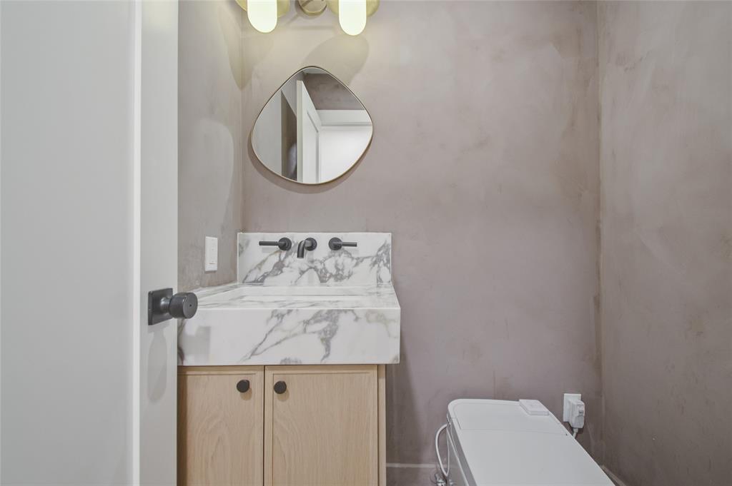 Stone-top vanity with a light wood-finish cabinet, matte black wall-mounted faucet, custom-shaped mirror, and smooth-finish walls