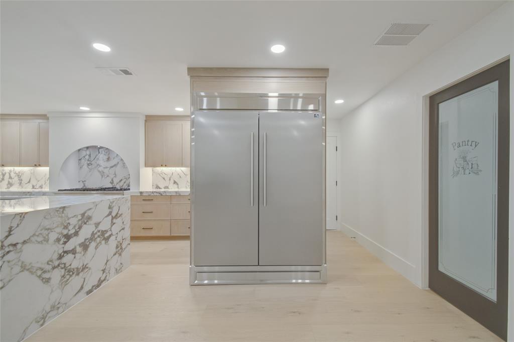 Custom cabinetry with a light wood finish, full-height built-in stainless steel refrigerator, stone countertops, integrated stone backsplash with arched detailing, and a door featuring a frosted glass panel with pantry signage