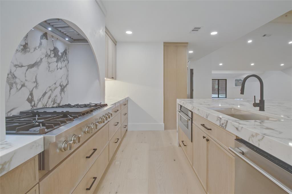 Kitchen featuring light wood-finish cabinetry, a striking arched cooktop alcove with a patterned stone backsplash, expansive stone countertops, a stainless steel gas range, and a matte black gooseneck faucet