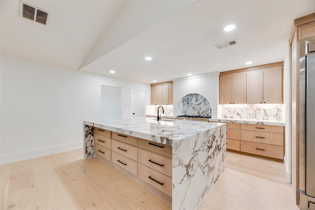 Kitchen featuring a waterfall-edge island with stone-finish countertops, custom light-wood cabinetry, recessed lighting, arched stove backsplash, and wide-plank wood-finish flooring