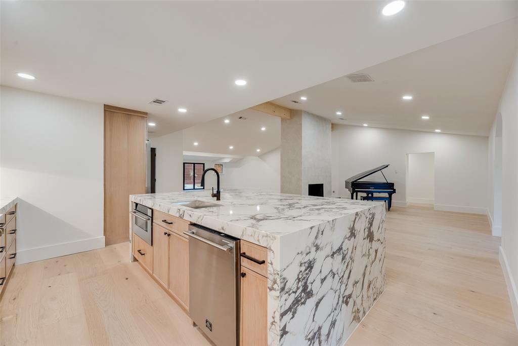 Kitchen featuring a waterfall-edge island with stone countertop, light wood-finish cabinetry, stainless steel dishwasher, matte black faucet, and recessed lighting