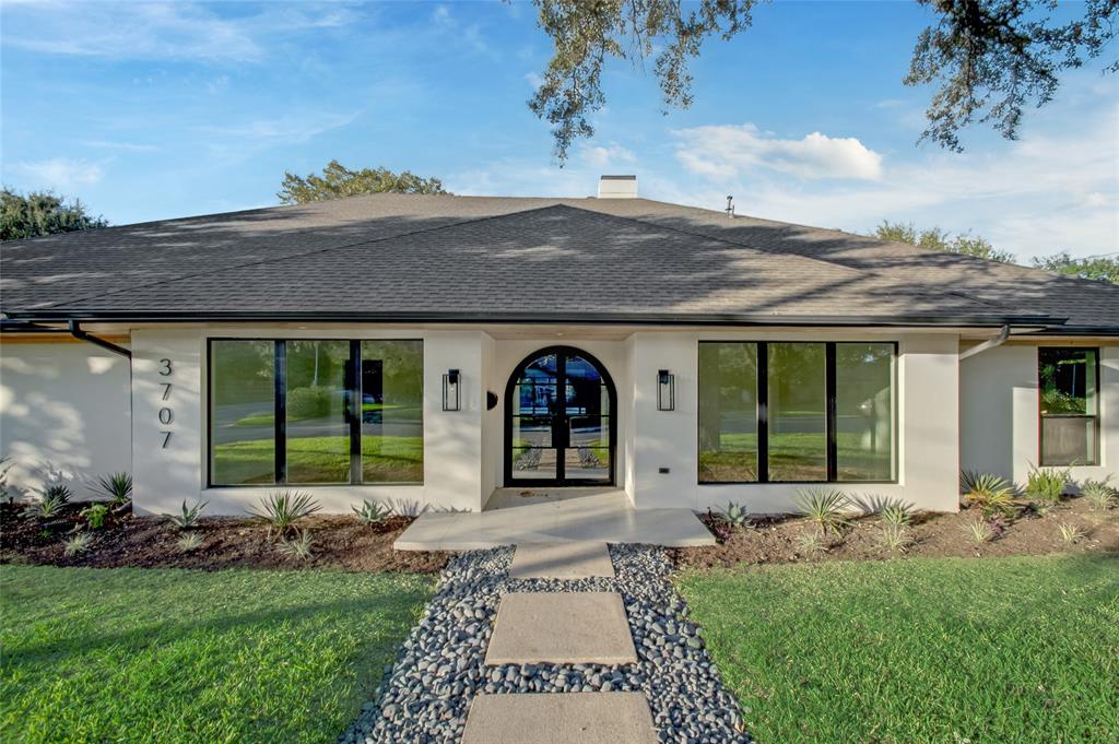 Single-story exterior featuring a light-toned stucco façade, an arched iron-framed entryway, large-pane picture windows, a multi-gabled roofline, and a concrete paver walkway bordered by decorative stone