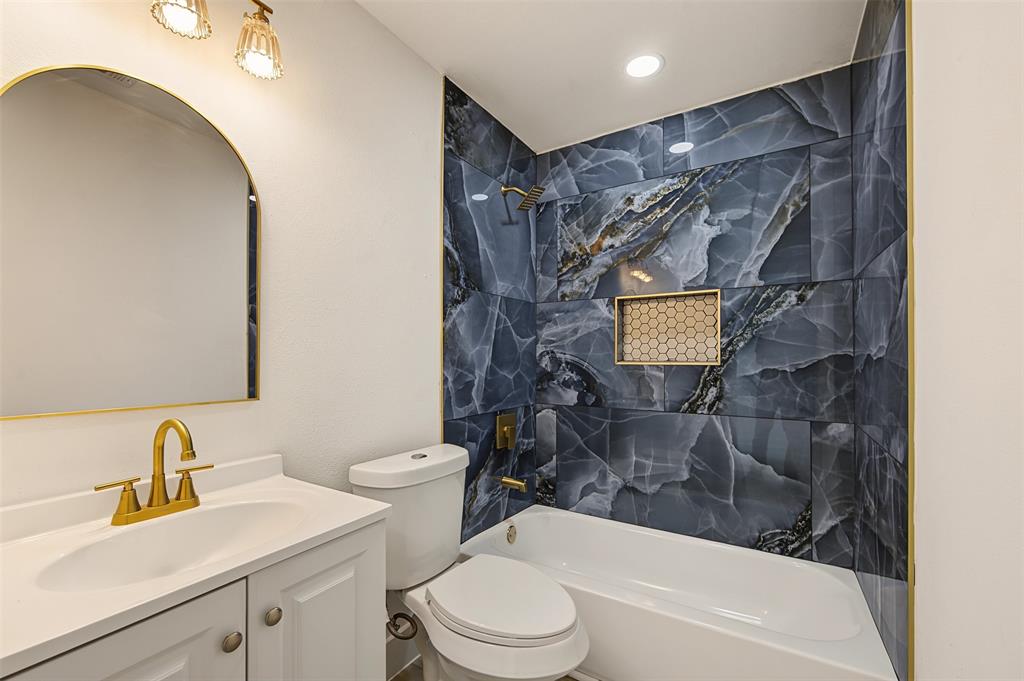 Bathroom featuring a white vanity cabinet with an integrated sink, arched mirror, brushed gold-tone hardware, and a shower-tub combination finished with large-format blue decorative tile and a hexagonal inset niche