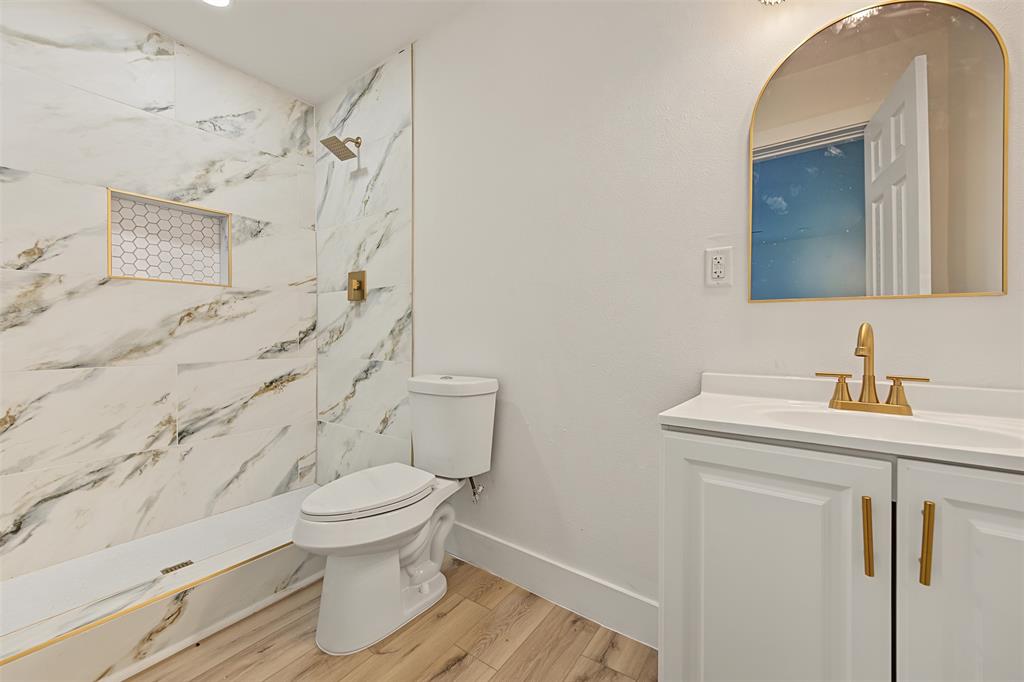 Bathroom featuring floor-to-ceiling veined tile, wood-finish flooring, a vanity with gold-toned fixtures, a built-in shower niche with hexagon tile, and an arched mirror