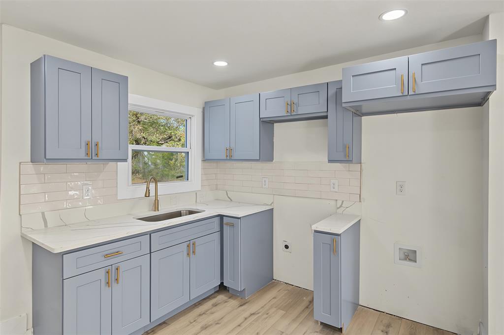 Kitchen featuring blue shaker-style cabinetry, light-toned stone-finish countertops, subway tile backsplash, brass-finish hardware, and wood-finish flooring