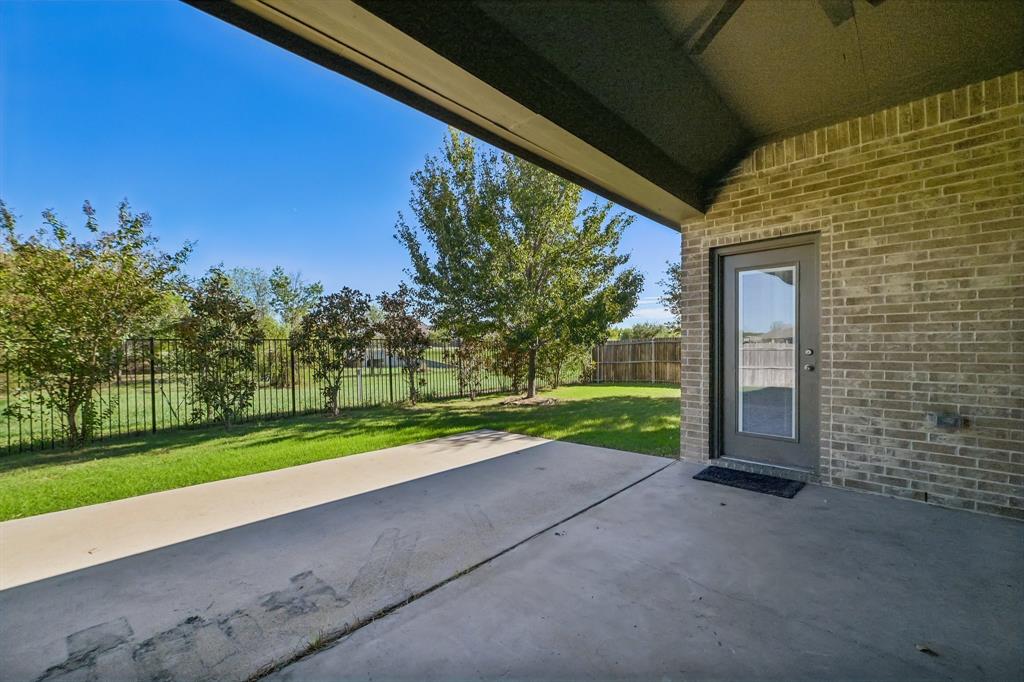Covered concrete patio featuring brick exterior siding, a full-glass inset door, metal perimeter fencing, and mature landscaping