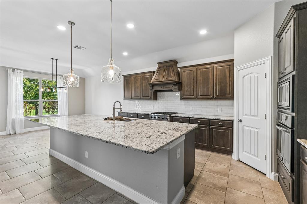 Kitchen featuring dark wood cabinetry, expansive stone-surface center island, built-in wall oven and microwave, stone-finish tile flooring, and decorative glass pendant lighting