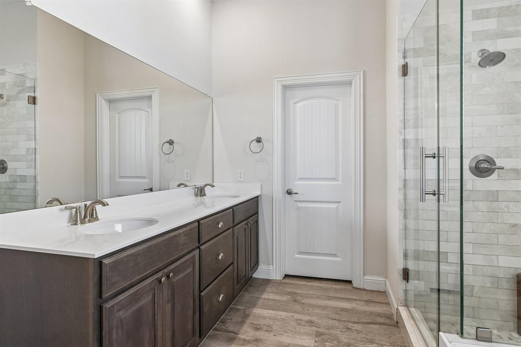 Dual-sink vanity with a solid-surface countertop, full-width wall mirror, wood-finish flooring, and a glass-enclosed shower featuring stone-style wall tile