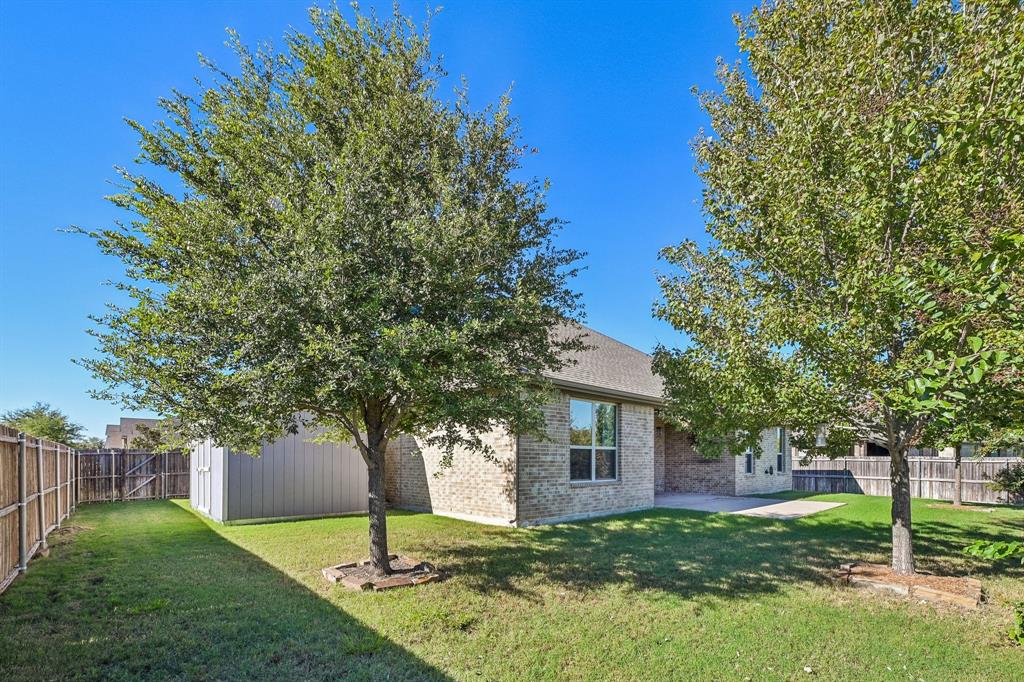 Rear exterior featuring brick siding, a concrete patio, mature trees, a manicured lawn, and wood-panel fencing