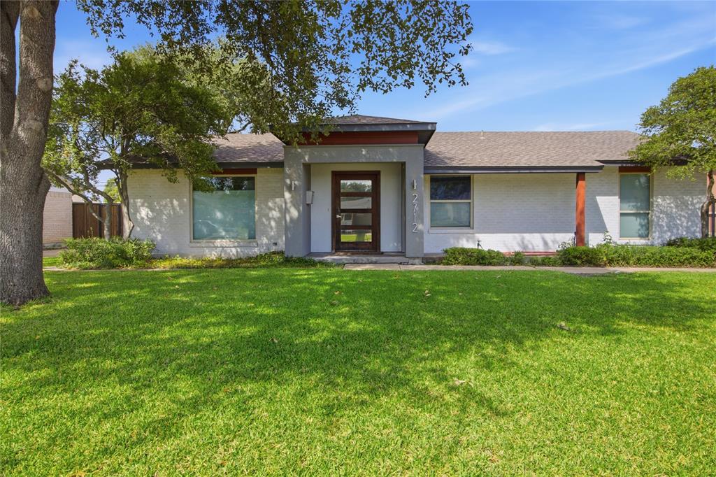 The daytime view reveals the home's welcoming curb appeal, with a spacious front lawn shaded by mature trees and established landscaping framing the exterior. The distinctive recessed entry, contemporary front door, and contrasting architectural accents add character, while the single-story layout creates a clean, inviting profile.