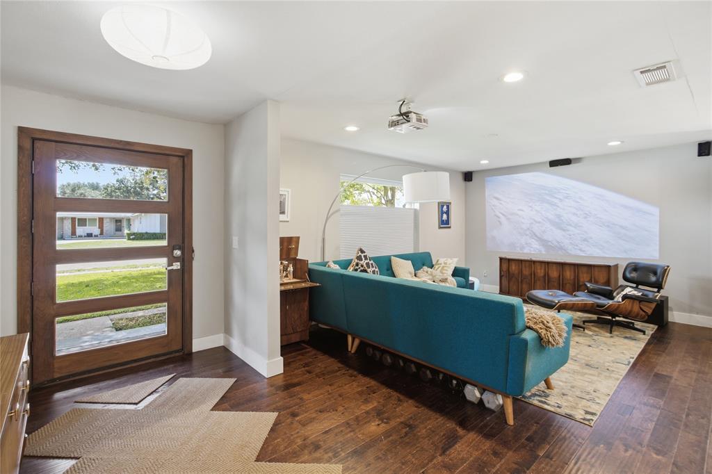 Step inside and discover a welcoming entry that opens into a stylish living room with mid-century modern influences. Rich, warm-toned flooring, clean architectural lines, and abundant natural light create an inviting atmosphere. The living area features a ceiling-mounted projector and speakers, creating a home theater experience for movie nights, sporting events, or relaxing at home. The combination of modern technology and comfortable living makes this a space designed to be enjoyed.