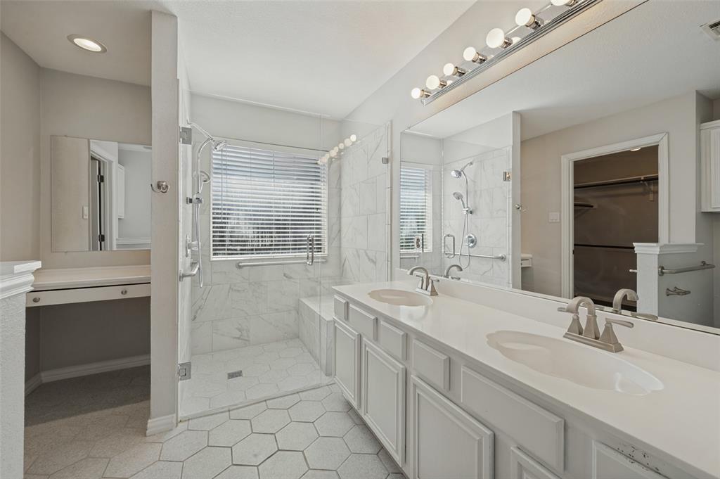 Dual-sink vanity with white cabinetry and solid-surface countertops, walk-in shower with integrated bench and wall-mounted spray fixture, hexagon-patterned tile flooring, and large-format wall tiling
