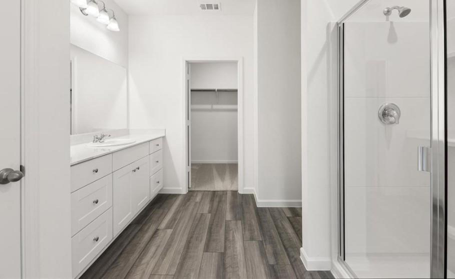 Bathroom featuring a white shaker-style vanity with stone-look countertop, wood-finish flooring, glass-enclosed shower, and built-in walk-in closet shelving