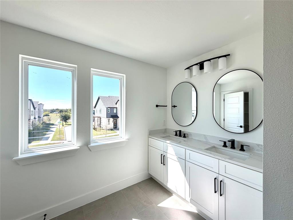 Dual-vanity cabinet with white shaker-style doors, light-toned stone countertop, dual undermount sinks, matte black hardware, and two circular mirrors
