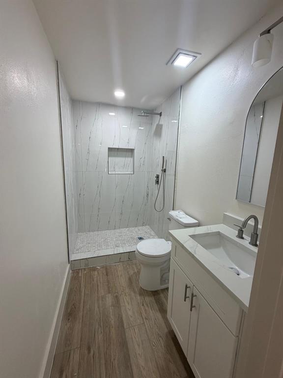 Bathroom featuring wood-finish flooring, white vanity with stone-style countertop, arched mirror, and walk-in shower with stone-look wall tiles, built-in recessed shelf, and pebble-style tile shower floor