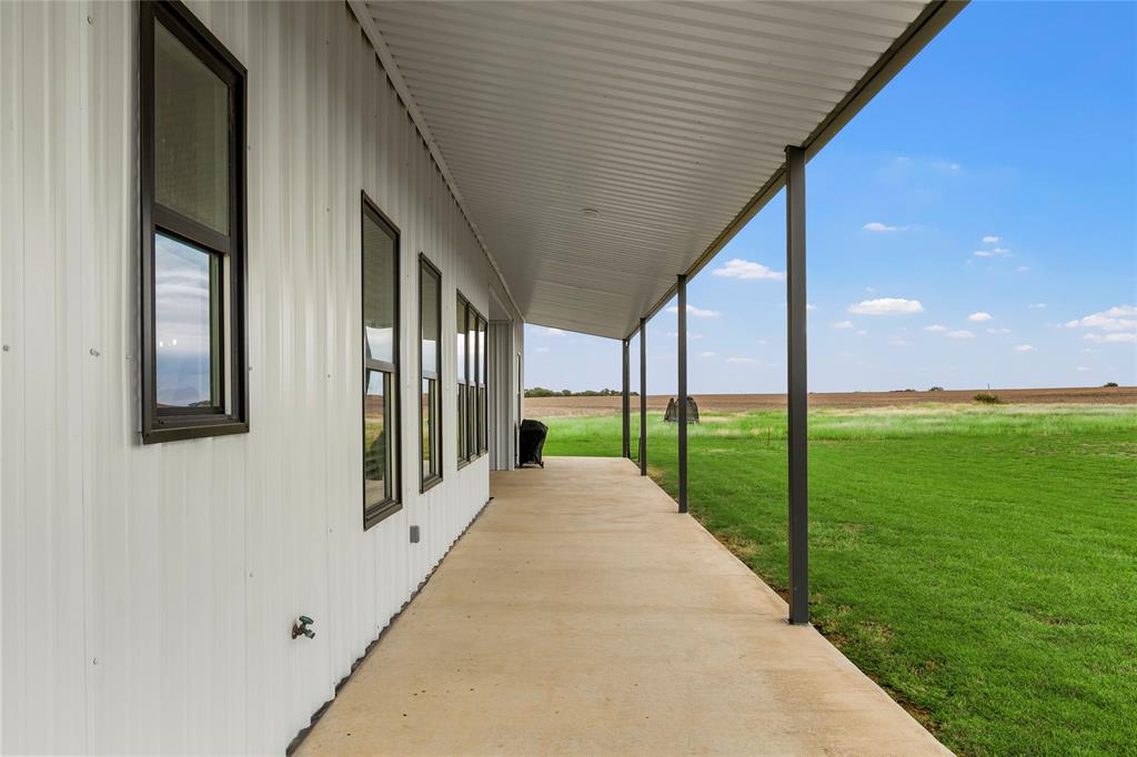 Covered concrete patio featuring a metal exterior wall, dark-framed windows, and metal structural supports overlooking an expansive open field